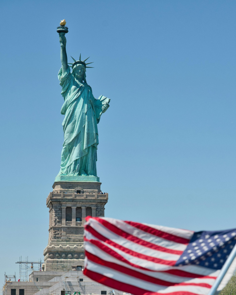 The Statue of Liberty on its pedestal against a clear sky, a US flag out of focus in the foreground