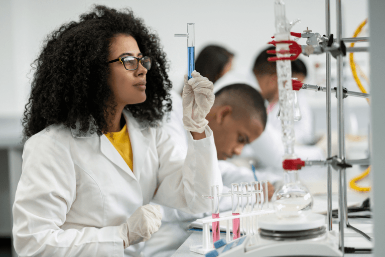 Researcher in a white lab coat holding up a test tube of blue liquid, with colleagues working behind her